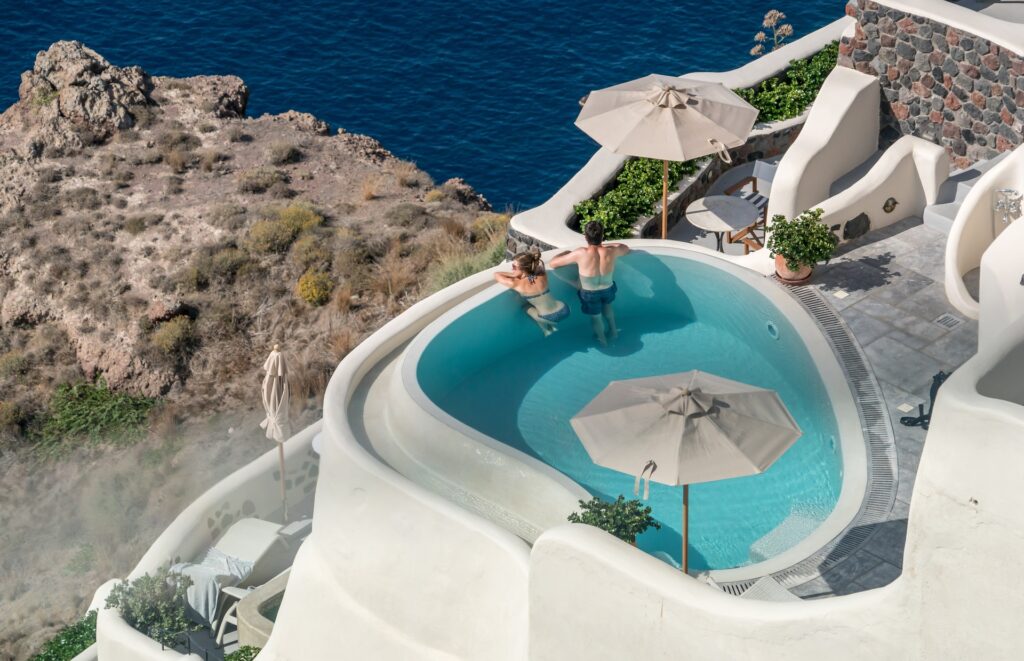 A romantic couple enjoying a dip in an infinity pool with a breathtaking view of Santorini's iconic shoreline and whitewashed buildings.