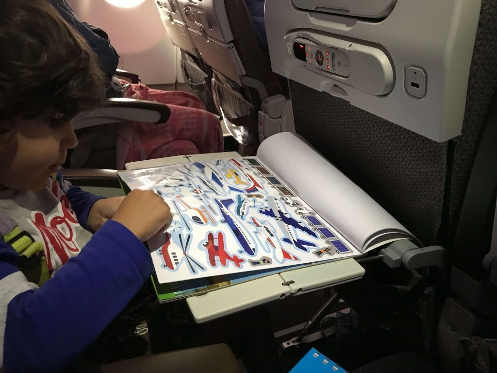 A young boy intently flipping through his sticker album while seated on an airplane.